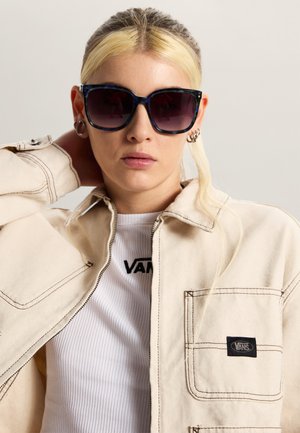 Ung kvinde med blondt hår iført mørke solbriller, en cremefarvet jakke med Vans-logo og en hvid ribbet Vans-t-shirt, poserer med hånden bag hovedet.