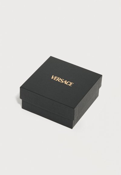 Μαύρο τετράγωνο κουτί με υφή και χρυσό λογότυπο «VERSACE» στο καπάκι, τοποθετημένο σε λευκή απλή επιφάνεια.