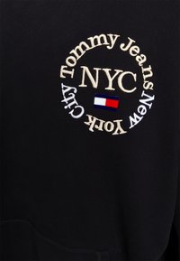 Svart hoodie med broderad kräm- och vit text "Tommy Jeans New York City" och en liten röd, vit och blå logotyp.