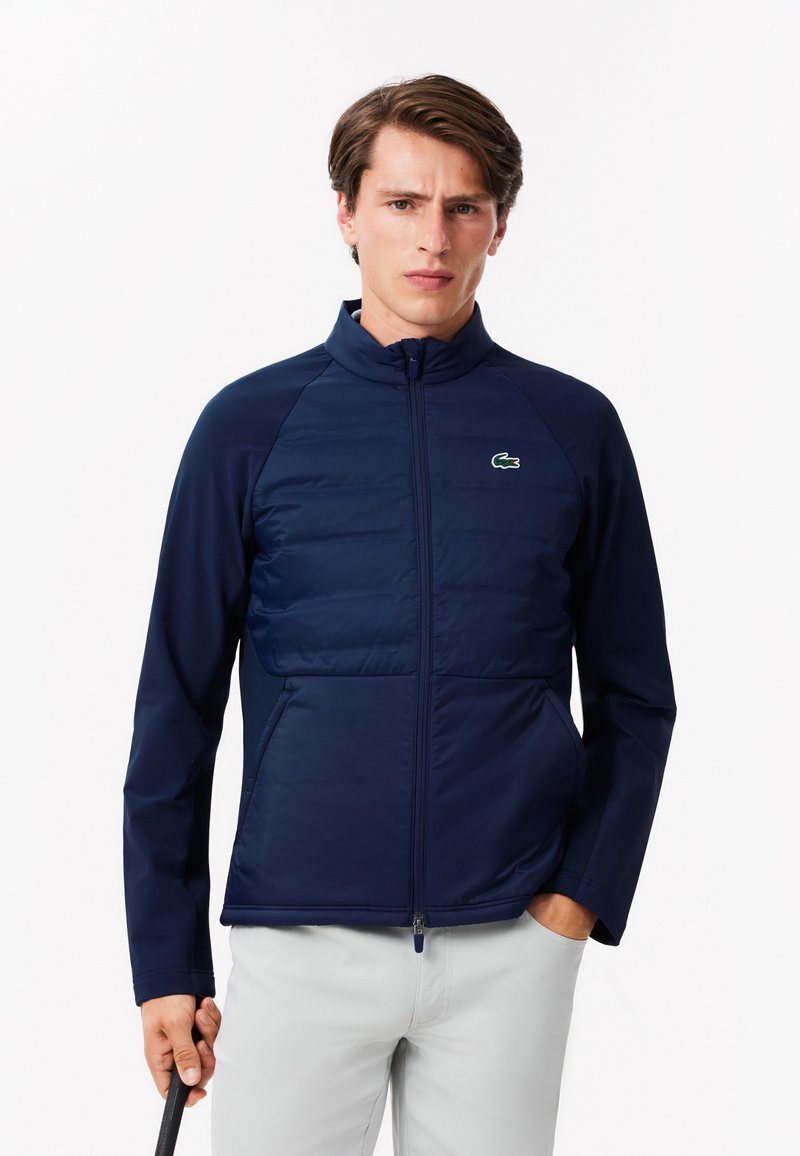 Giacca blu navy con zip frontale, parte superiore trapuntata e parte inferiore liscia. Presenta un colletto alto e il logo Lacoste sul petto.