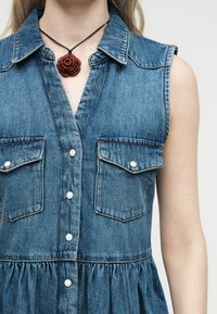 Μπλε τζιν φόρεμα χωρίς μανίκια με κουμπιά στη μέση και τσέπες με καπάκι, συνδυασμένο με μενταγιόν σε σχήμα σκοτεινού τριαντάφυλλου σε μαύρο κορδόνι.