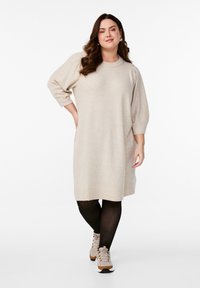 Beige oversized tröja klänning med trekvartsärmar och ribbade manschetter, tillsammans med svarta strumpbyxor och beige sneakers. Slät stickad textur.