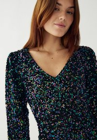 Juoda blizgučiais siuvinėta suknelė su V formos iškirpte ir ilgomis rankovėmis. Suknelė turi daugiaspalvius atspindinčius blizgučius, sukuriantį tekstūruotą paviršių.