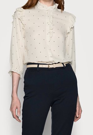 Kvinde iført en hvid prikket bluse med flæser og mørkeblå bukser, accessoriseret med et lyst beige bælte.