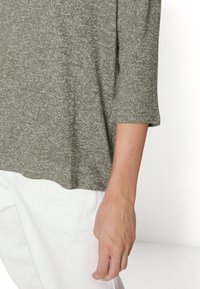 Haut gris texturé à manches trois-quarts, coupe décontractée, associé à un pantalon blanc ; mise en avant de la douceur du tissu et du style casual.