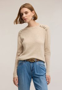 Beige pletený svetr s dekorativními cvočky na ramenou, doplněný modrými džíny a hnědým opaskem, na neutrálním pozadí.