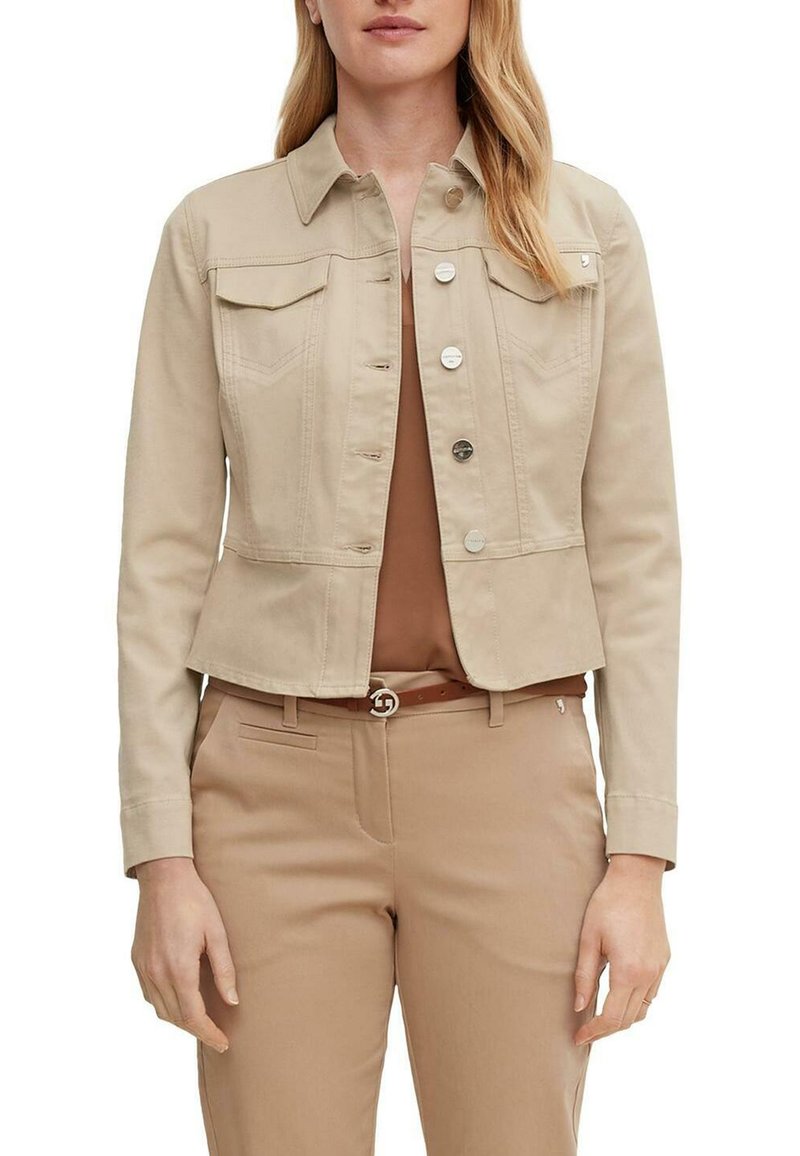 Femme portant une veste beige boutonnée par-dessus un haut marron et un pantalon beige avec une ceinture marron sur fond blanc.