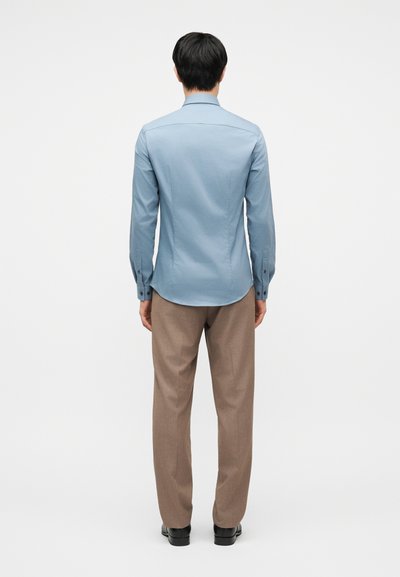 Άνδρας που στέκεται πλάτη, φοράει ανοιχτό μπλε εφαρμοστή μακρυμάνικη πουκάμισο, καφέ παντελόνι και μαύρα παπούτσια, σε απλό λευκό φόντο.