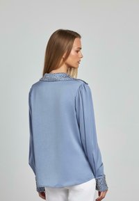 Blusa de manga larga azul en tejido satinada con cuello y puños adornados, que presenta una textura suave y un brillo sutil.