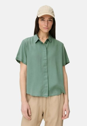 Femme portant une chemise vert clair à manches courtes boutonnée, un pantalon beige et une casquette de baseball beige, debout devant un fond blanc uni.
