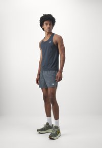 Débardeur de sport gris foncé, associé à un short gris. Chaussures de sport avec des accents verts et jaunes. Le modèle se tient sur un fond neutre.
