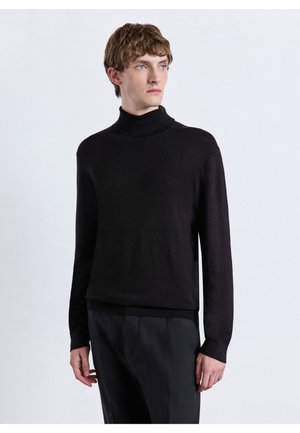 Giovane uomo con maglione dolcevita nero e pantaloni scuri, in piedi con le mani rilassate, guarda di lato su uno sfondo chiaro e uniforme.