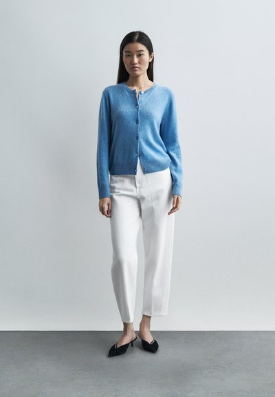 Γυναίκα που φοράει μπλε ζακέτα με κουμπιά, λευκό ψηλόμεσο παντελόνι και μαύρες μυτερές γόβες με λουράκι στη φτέρνα, στέκεται σε γκρι πάτωμα.