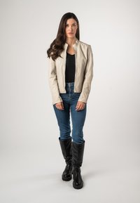 Lichtbeige leren jas, rits aan de voorkant, ronde hals, gecombineerd met donkere denim jeans en kniehoge zwarte laarzen. Eenvoudig, modern ontwerp, gestructureerd oppervlak.