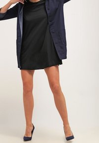 Svart klänning med lös passform, i kombination med en marinblå knapp-up jacka. Modellen bär marinblå klackar. Bakgrunden är helt vit.