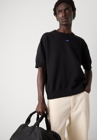 Pulover scurt din tricotaj negru, cu guler rib și mâneci scurte, purtat cu pantaloni de culoare crem, ținând o geantă sport neagră.