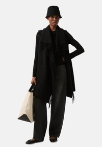 Sort strikket poncho med frynsekant, kombineret med højttaljede mørkegrå jeans og sorte balletsko. Bærer en beige lærredsnettaske.