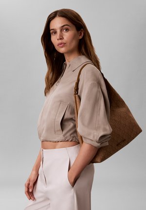 Pikkade pruuni juustega naine kannab beežikat lühendatud jope ja heledaid pükse, kantakse suurt pruuni mustrilist õlakotti, seisab halli tausta ees.