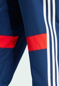 Granatowe spodnie sportowe z czerwonymi poziomymi paskami. Mają trzy białe pionowe paski po boku. Gładki, lekki materiał.