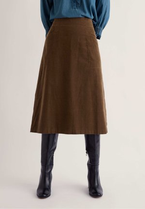 Femme portant une jupe midi marron associée à des bottes noires hautes jusqu'aux genoux et un chemisier bleu à manches longues, debout devant un fond uni.