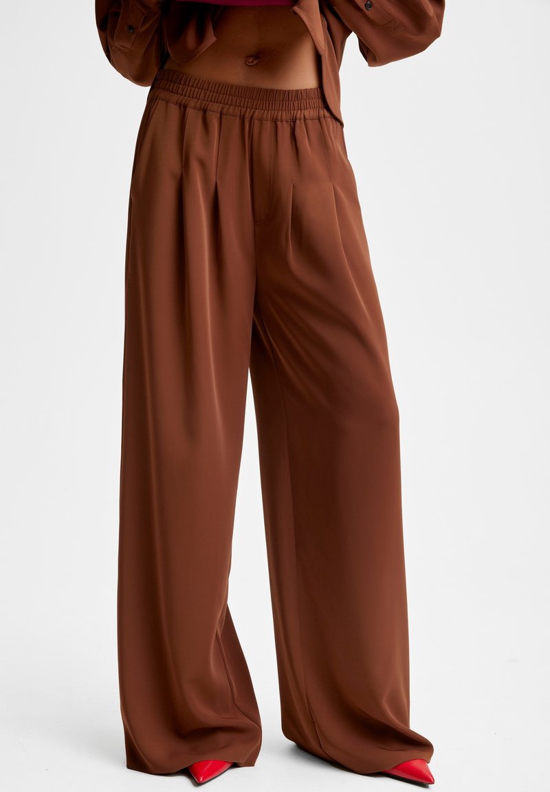 Pantalon marron à jambes larges avec taille élastique porté par une personne, associé à des chaussures rouges pointues sur un fond uni.
