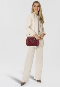 Rote gesteppten Handtasche mit Kettenakzenten und einem dreieckigen Logo, in der Hand gehalten. Das Model trägt einen cremefarbenen Anzug und beigefarbene Absätze vor einem schlichten Hintergrund.