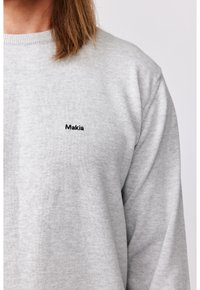 Personne portant un sweat-shirt gris clair avec le mot « Makia » brodé en petites lettres noires sur le côté gauche de la poitrine, montrant une partie de l'épaule et du cou.
