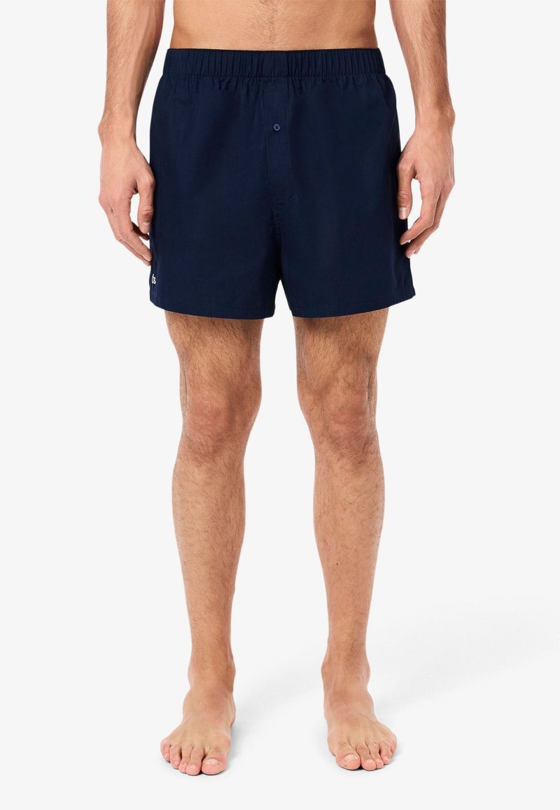 Navy blue swim trunks with an elastic waistband and a single button, featuring a small logo on the left side. Smooth, lightweight fabric.
