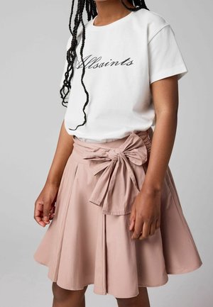 Ung kvinde med fletninger iført en hvid AllSaints t-shirt, stukket ned i et blommefarvet rosa skørt med en stor bundet sløjfe i taljen.
