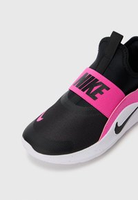 Baskets slip-on Nike noires et roses avec tige en maille, sangle rose portant l'étiquette "NIKE", semelle blanche et logo swoosh noir sur le côté et sur le bout.