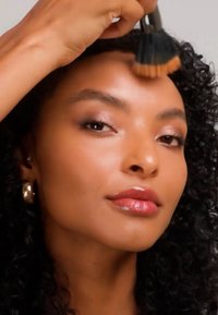 Femme aux cheveux bouclés appliquant du fond de teint sur le front avec un grand pinceau, portant des boucles d'oreilles dorées en forme d'anneau.