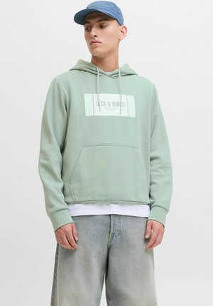 Sudadera verde menta con un bolsillo frontal, capucha con cordones y parche de logotipo blanco. Combinada con jeans azul claro y una gorra azul.