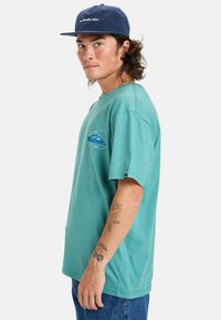 Camiseta de algodón turquesa con un ajuste relajado, que presenta un gráfico de logo azul en el pecho. Mangas cortas y costuras visibles. Diseño casual.