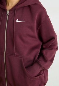 Moleton com fecho éclair numa cor bordô, feito de um tecido suave, com um bolso à frente, punhos canelados e um logo branco da Nike no lado esquerdo do peito.