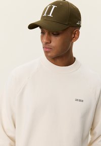 Άνδρας που φοράει πράσινο-ελαιί καπέλο με λευκά γράμματα "II" και κρεμ φούτερ με το κείμενο "LES DEUX", κοιτάζοντας προς τα κάτω στο πλάι.