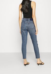 Mujer con cabello largo y oscuro, vistiendo una blusa negra sin mangas, jeans ajustados de tiro alto azul con dobladillos desflecados y sandalias de tacón negras.