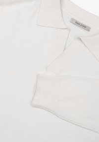 Polo shirt bianco, in cotone, colletto piatto, maniche lunghe con polsini a costine, design minimalista, con un piccolo etichetta del marchio sul colletto.