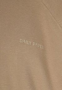 Tessuto a coste beige con una finitura testurizzata. Logo "DAILY PAPER" in lettere in rilievo posizionato sul lato sinistro inferiore.
