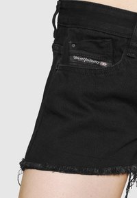 Mustat farkkushortsit, joissa on revityt reuna, taka-taskussa Diesel Industry -merkki, esiteltynä yksinkertaisen taustan edessä.