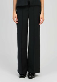 Femme portant un pantalon large noir et des chaussures en cuir verni noires, se tenant avec les mains détendues le long du corps.