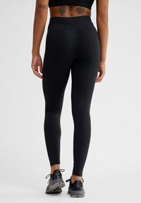 Schwarze Leggings mit hoher Taille aus Stretchstoff, mit glatter Textur und figurbetontem Schnitt. Getragen mit schwarzen Sportschuhen.