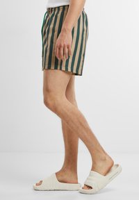Gestreifte Badeshorts mit vertikalen blauen und beigen Streifen, elastischem Bund mit Kordelzug, kombiniert mit weißen Slidesandalen.