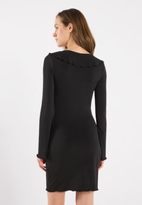 Svart kjole med lange ermer, volangkant ved halsen, tettsittende design og myk tekstur, som rekker til midt på låret. Glatt stoff og elegant utseende.