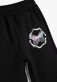 Zwarte trainingsbroek van zacht materiaal, met een elastische tailleband en een afbeelding van een gestileerde vlinder met omringende tekst.