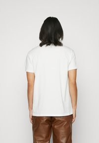 T-shirt blanc à manches courtes avec un col rond, en coton, vu de dos, associé à un pantalon en cuir marron.