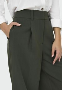 Grønne høytlivsbukser med folder, har sidelommer og glatt tekstur, kombinert med en hvit skjorte med knapper.