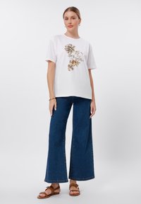 Hvit bomulls t-skjorte med blomstertrykk i gult og svart, kombinert med vide blå denimbukser og brune sandaler.