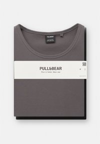 Débardeur gris avec une texture côtelée, plié sur un tissu noir, présentant une étiquette blanche avec le nom de la marque et un slogan.