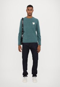 Teal knit sweater with a white "W" logo, dark jeans, and white sneakers. Model holds a black crossbody bag with a leather strap.
