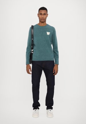 Teal knit sweater with a white "W" logo, dark jeans, and white sneakers. Model holds a black crossbody bag with a leather strap.